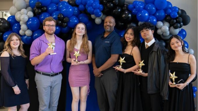 six honorees with their awards stand in front of a blue and black balloon arch with the college president at center