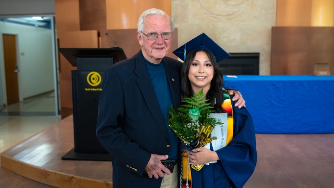 Trustee Trojan stands in a navy suit with his arm around Crystal who is pictured in her cap and gown